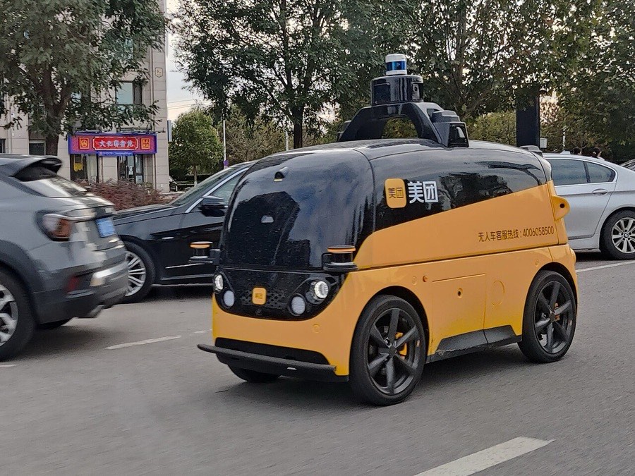 Ein fahrerloser Lieferwagen von Meituan im Straßenverkehr, gelb und schwarz lackiert, mit Lidar und Kameras auf dem Dach.