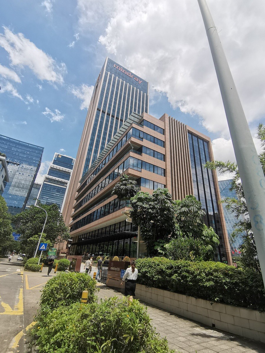 Der Hauptsitz von Mindray in Nanshan, Shenzhen: ein braun verkleideter Hochhausturm mit dem Firmennamen an der Fassade, davor Bäume und eine Zufahrt.